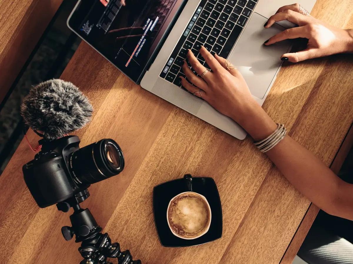Mãos digitando em um laptop, ao lado de uma câmera e uma xícara de café